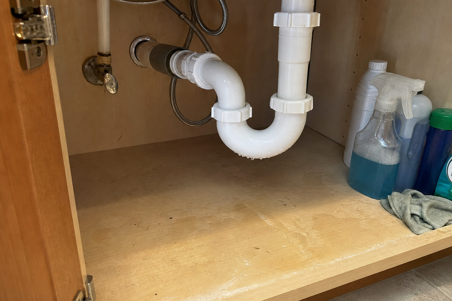Under-sink drain trap with water droplets and damp cabinet floor